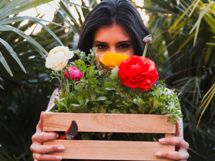 A woman smiles while holding a wooden box filled with colorful flowers.