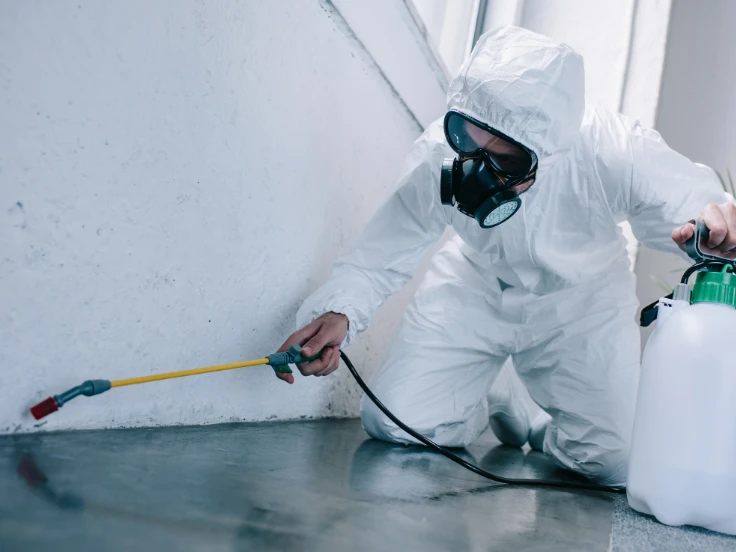 A man wearing a white protective suit is spraying a white substance, possibly for sanitation or pest control.