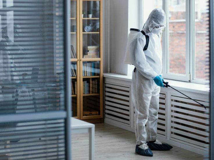 A man wearing a white suit and protective gear sprays a room, focusing on sanitizing surfaces for health safety.