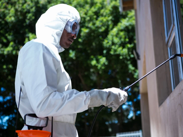 A man in a white protective suit is applying spray to a window, highlighting safety measures in his cleaning process.