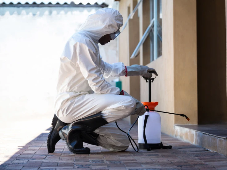 A man dressed in a white suit and protective gear uses a white hose to spray, ensuring safety while working.