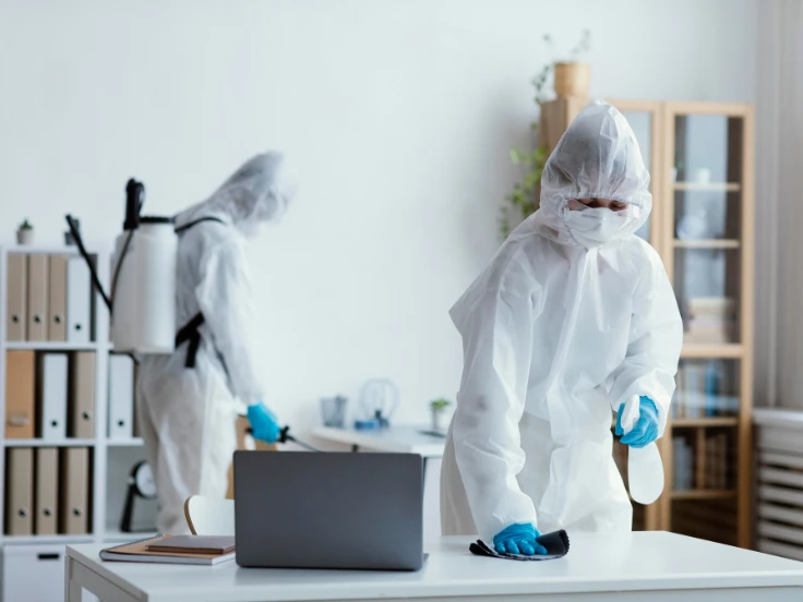 Two people wearing masks and protective clothing are intently working together on a laptop in a controlled setting.
