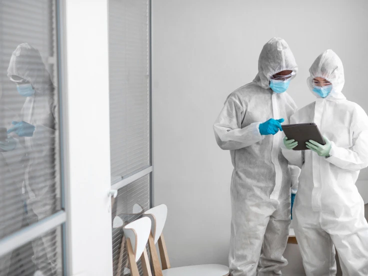 Two individuals in protective suits and masks stand by a computer, focused on their work in a safety-conscious environment.