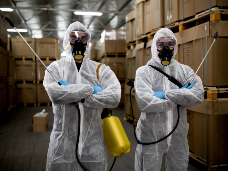 Two individuals in protective suits stand inside a spacious warehouse, surrounded by shelves and equipment.