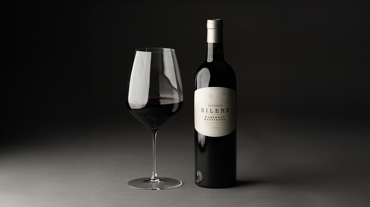 A bottle of Capensis Silene Cabernet Sauvignon wine standing next to a glass of red wine on a dark background.