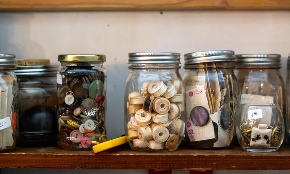 Collection of recycled glass jars neatly organized on a wooden shelf, holding sewing supplies, buttons, and small notions-Green Living Ideas