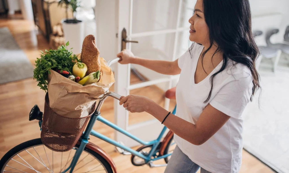 Person standing in front of an open refrigerator stocked with fresh produce while writing a grocery inventory list on paper